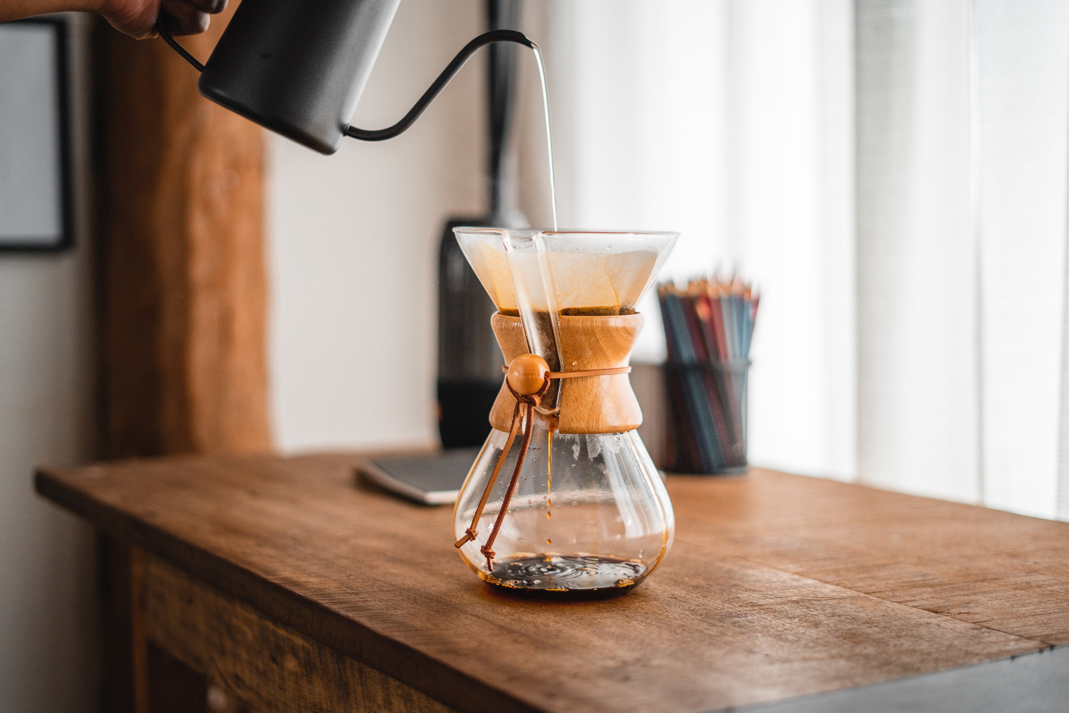 Black gooseneck kettle pouring over a glass Chemex on a wooden table in warm natural light.