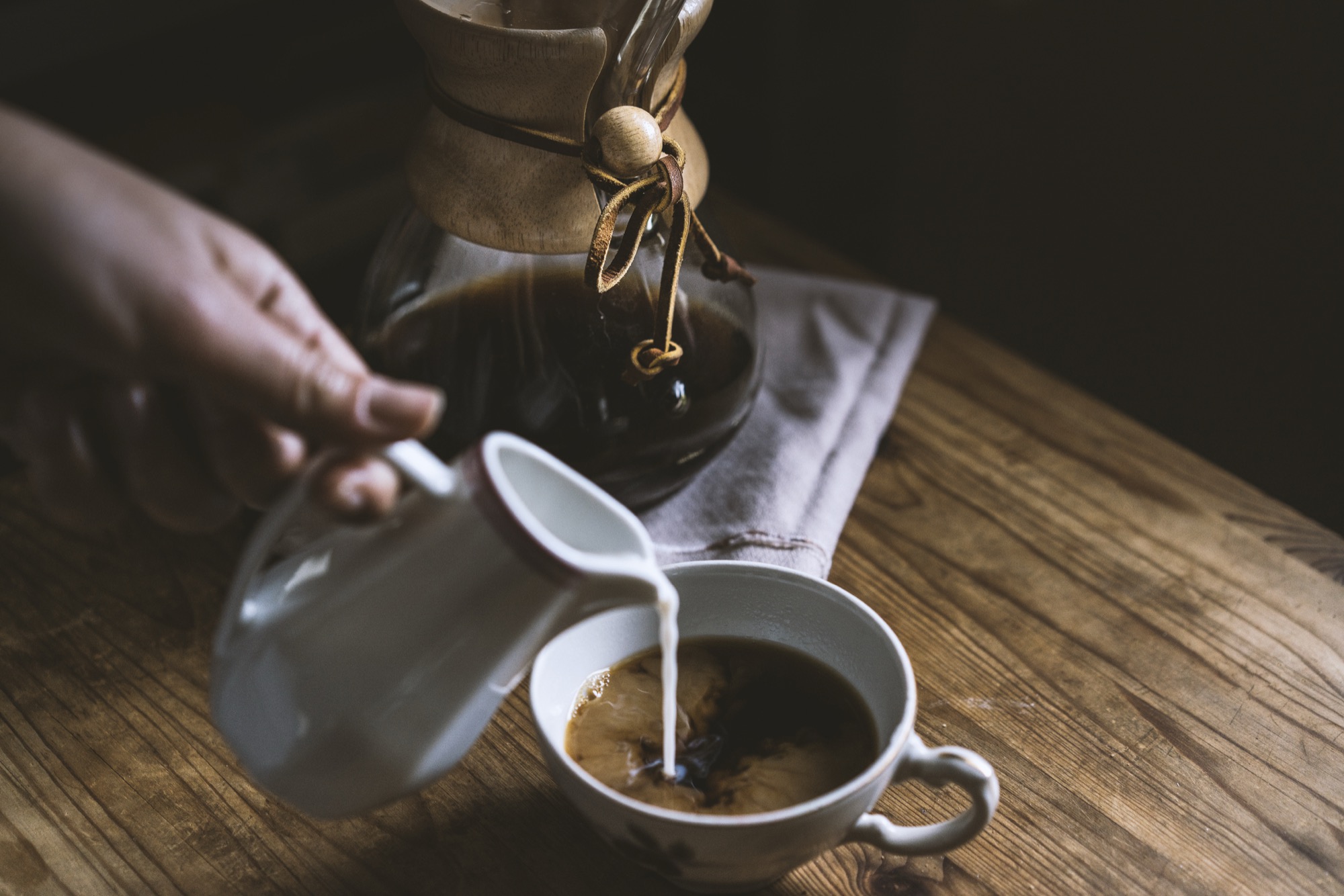 Milk being poured into a cup of coffee beside a Chemex on a wooden table.
